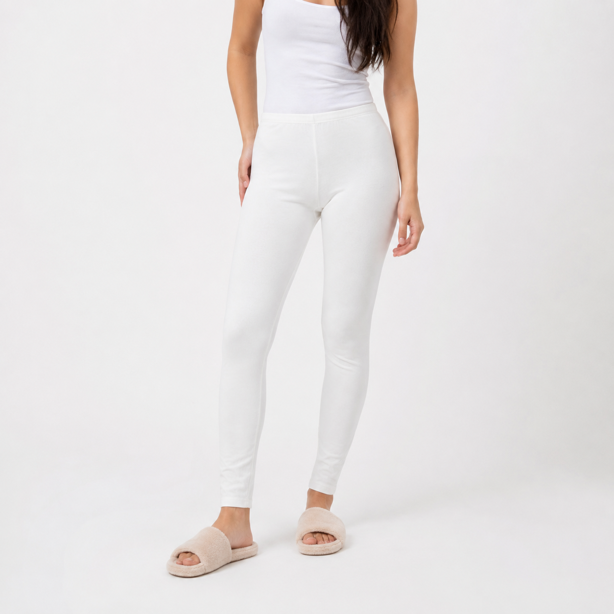 A model wearing white high-waisted leggings, a white tank top, and beige slide sandals, standing against a plain white studio background.
