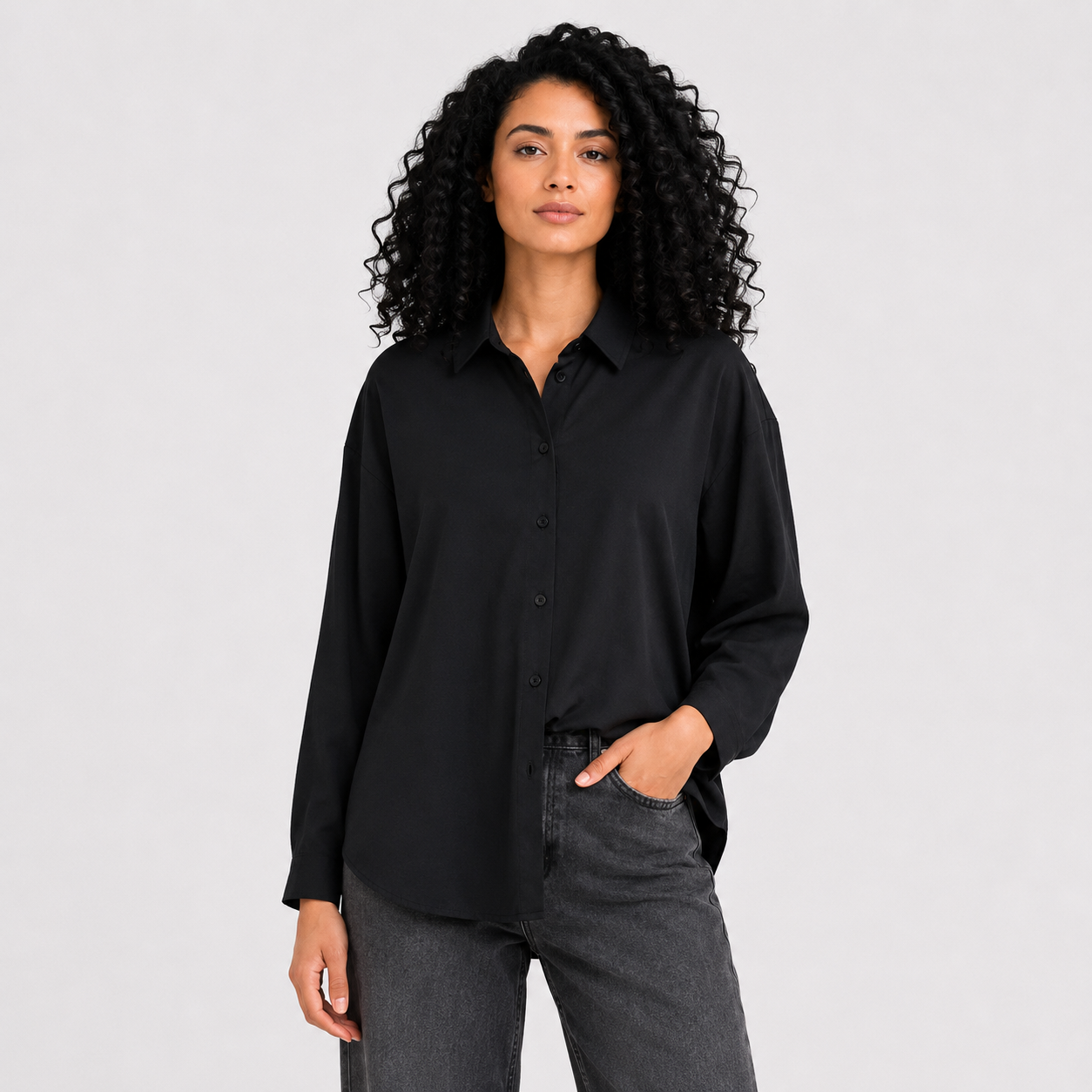 A woman with curly black hair wearing a black satin long-sleeve button-up shirt and dark gray jeans, standing against a light gray studio background.