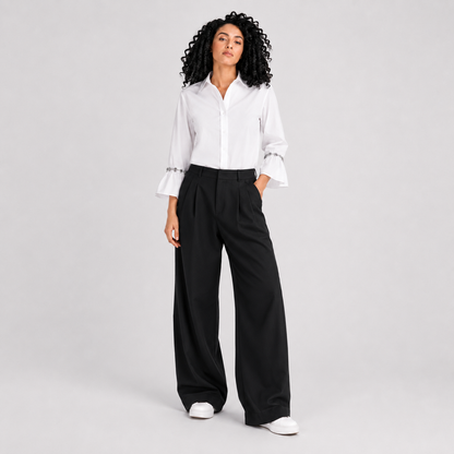 A model with curly black hair stands against a light gray studio background wearing a white long-sleeve button-up shirt with ruffled cuffs, black wide-leg pleated trousers, and white sneakers.
