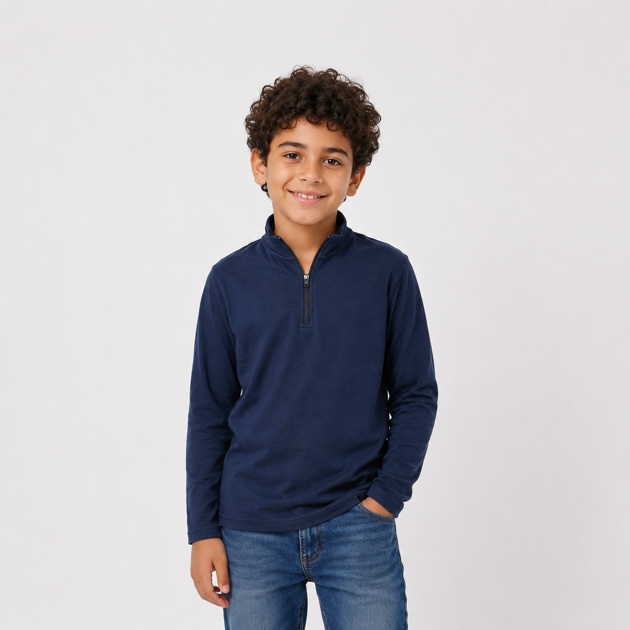 A young boy with curly brown hair wearing a navy blue long-sleeve quarter-zip pullover and blue jeans, standing against a light gray studio background.