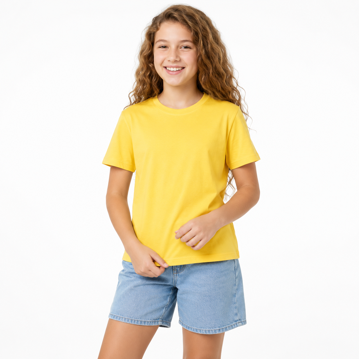A young girl with long curly brown hair smiling, wearing a solid yellow short-sleeve crewneck t-shirt and light blue denim shorts, standing against a plain white studio background.