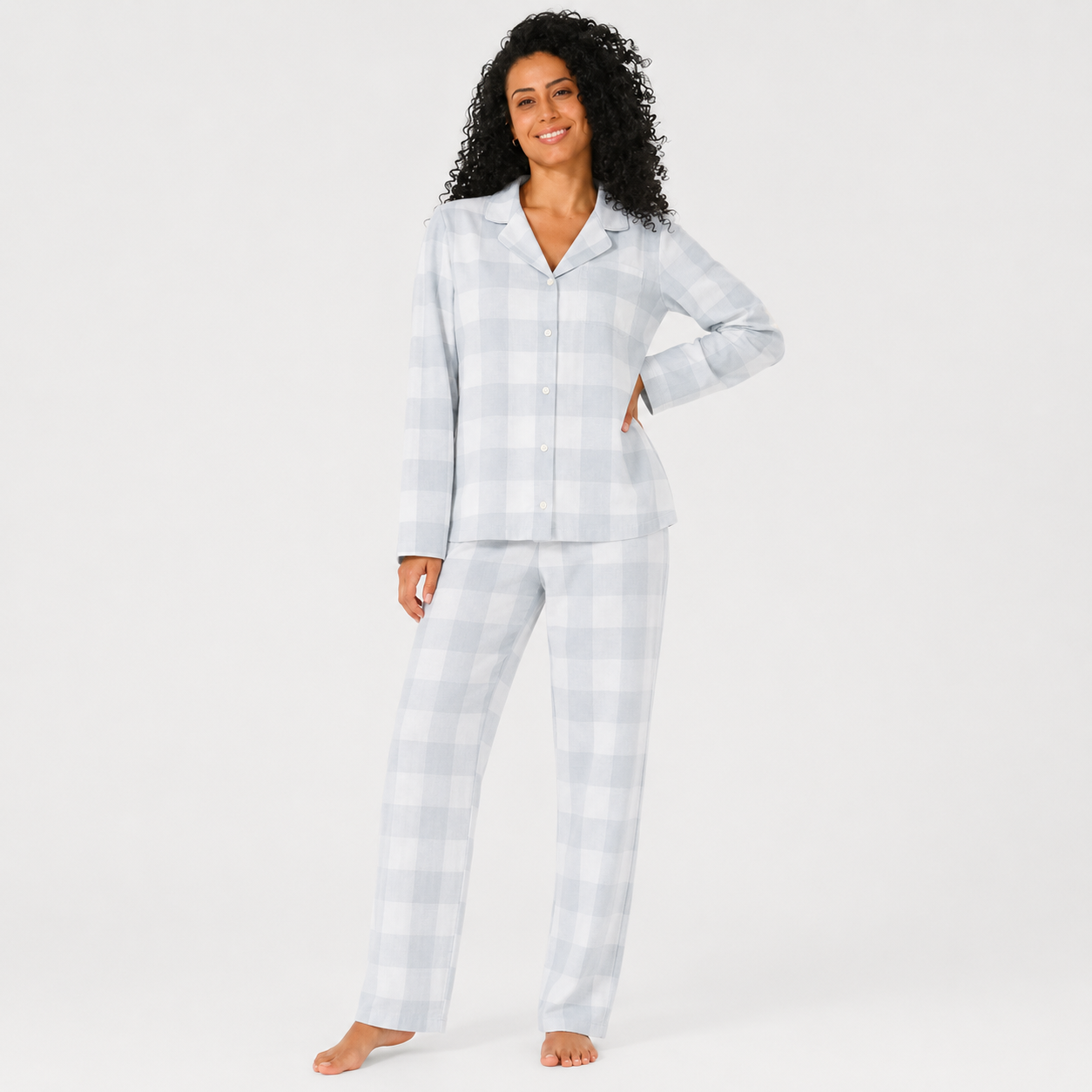 A woman modeling a light gray and white gingham check pajama set, consisting of a long-sleeve button-up top and matching pants, against a plain white studio background.