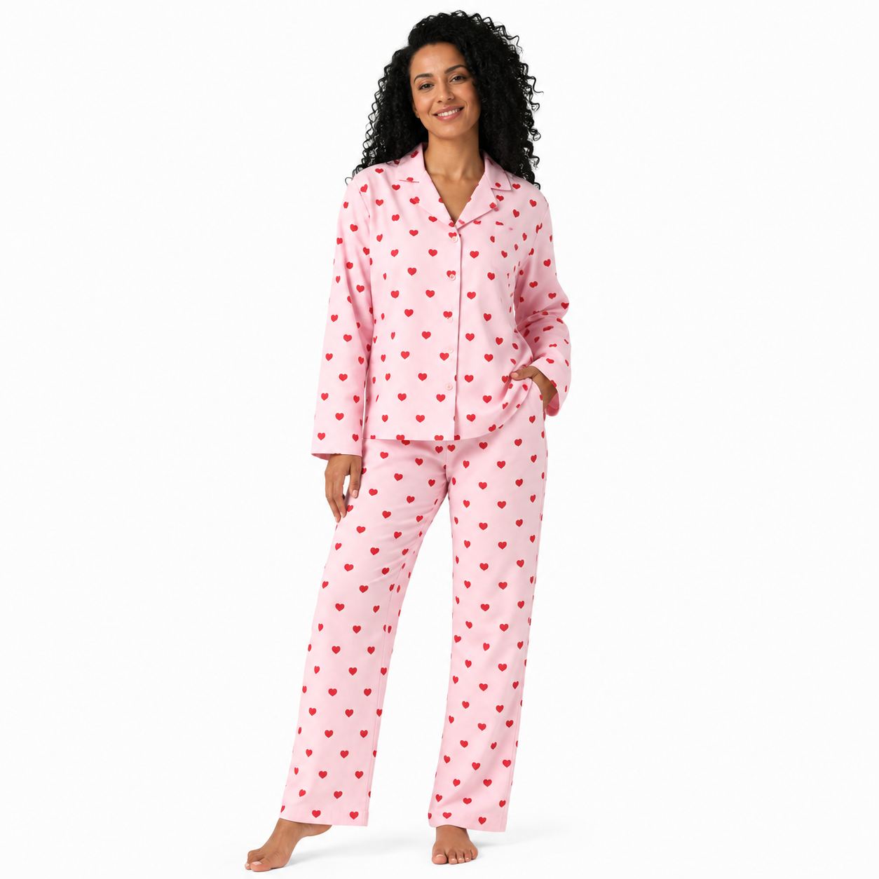 A woman modeling a two-piece pink pajama set with a red heart pattern, consisting of a long-sleeve button-up top and matching pants, against a plain white background.