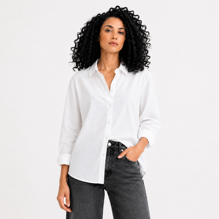 A woman with curly black hair wearing a white long-sleeve button-up shirt and dark gray jeans, standing against a plain white studio background.