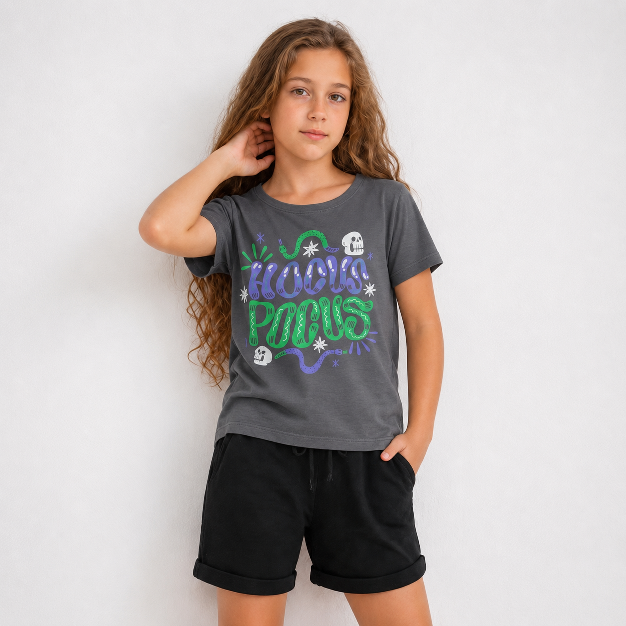 A young girl with long brown hair stands against a light gray studio background wearing a dark gray graphic t-shirt with 'Halloween Pocus' in green and purple text, paired with black shorts.