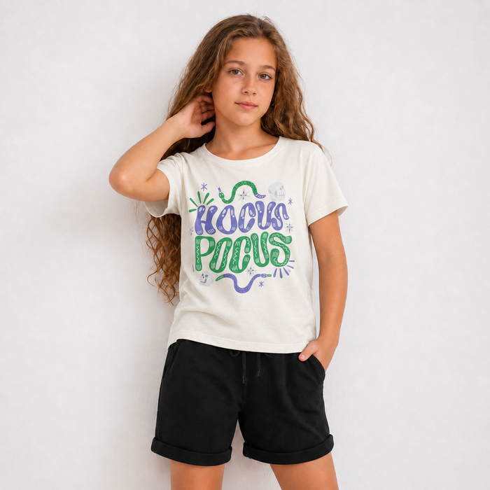 A young girl with long wavy brown hair stands against a light gray studio background wearing a cream-colored graphic t-shirt with 'KOOKS POCAUS' written in purple and green, paired with black shorts.