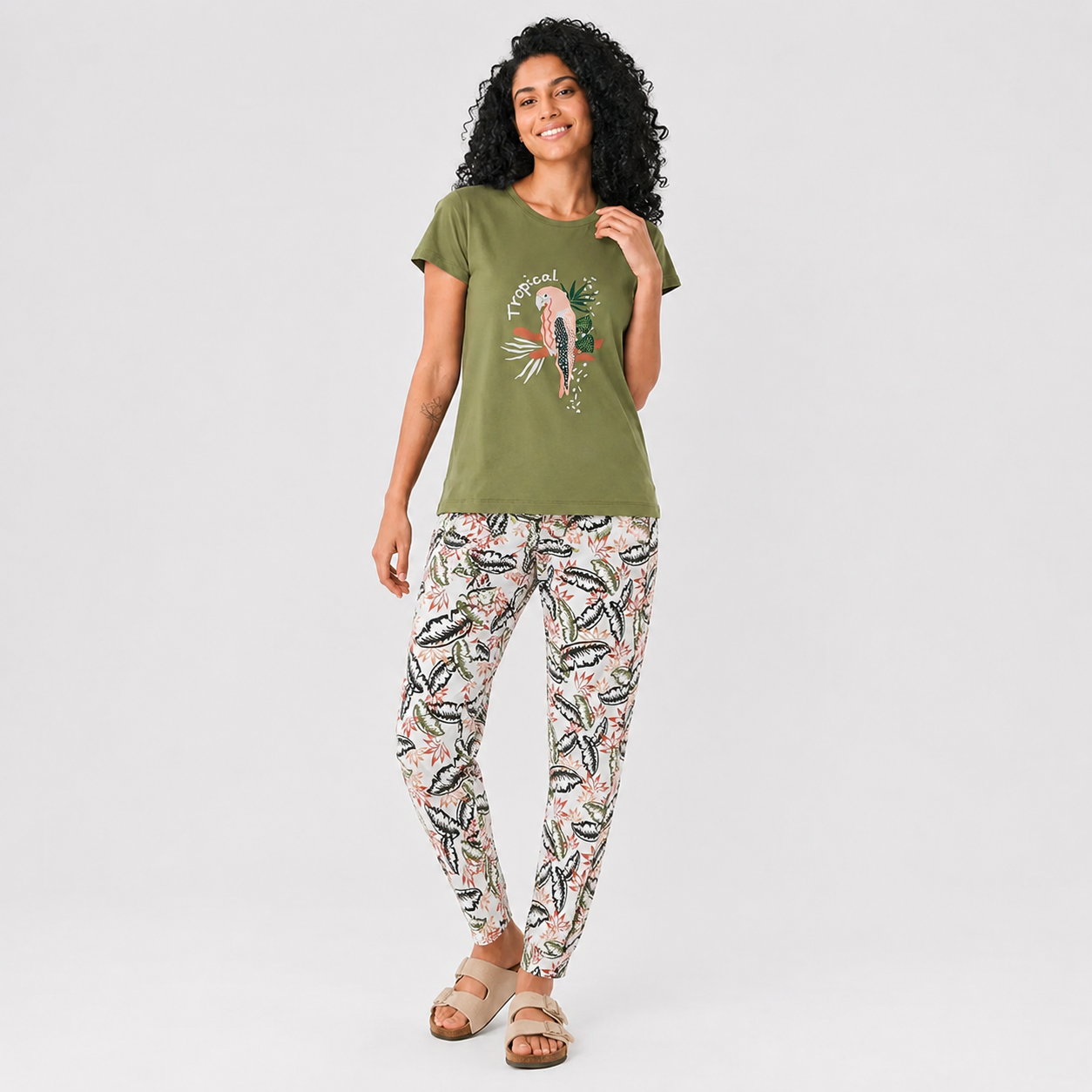 A woman modeling a two-piece lounge set consisting of an olive green t-shirt with a tropical parrot graphic and matching patterned pants with a palm leaf print, set against a plain light gray background.