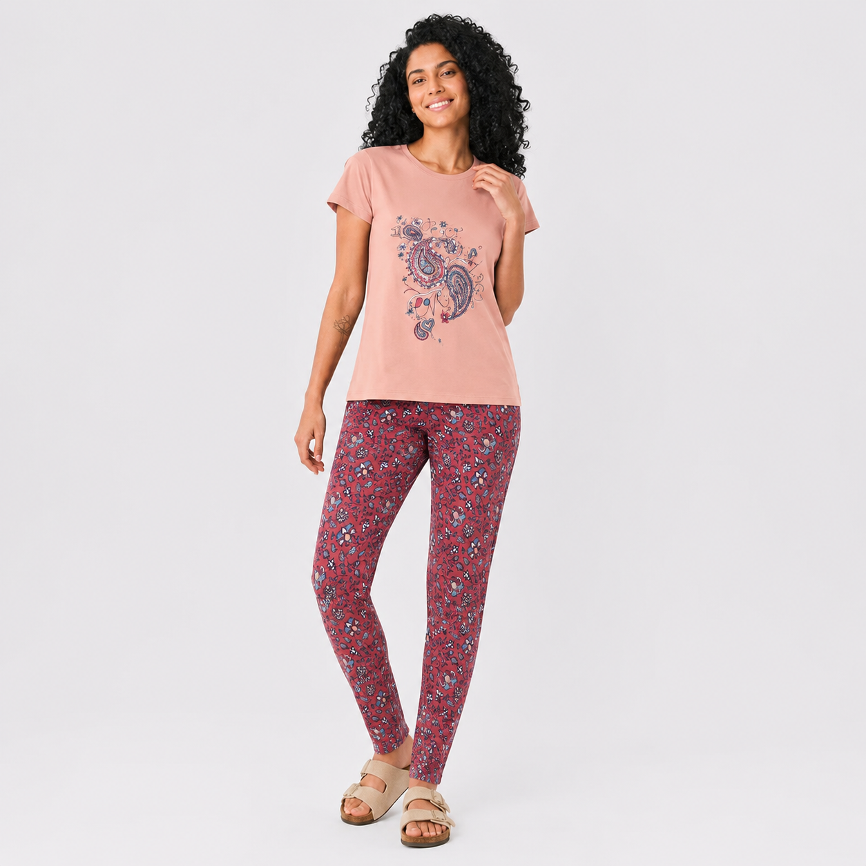 A woman modeling a two-piece loungewear set consisting of a dusty pink t-shirt with a paisley graphic and matching red floral print leggings, set against a plain light gray background.