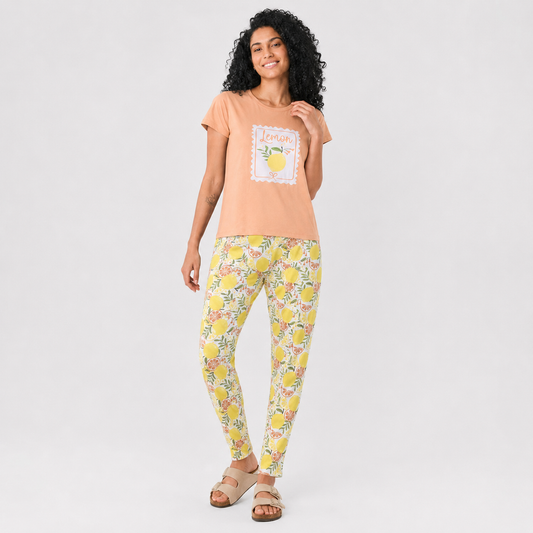 A woman modeling a peach short-sleeve t-shirt with a lemon graphic and yellow floral print leggings, standing against a light gray studio background.