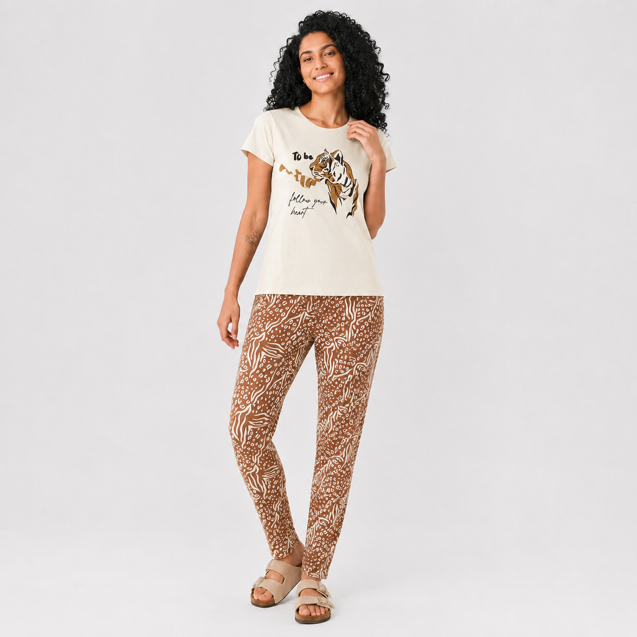A woman modeling a cream graphic t-shirt and brown leopard-print leggings against a light gray studio background.