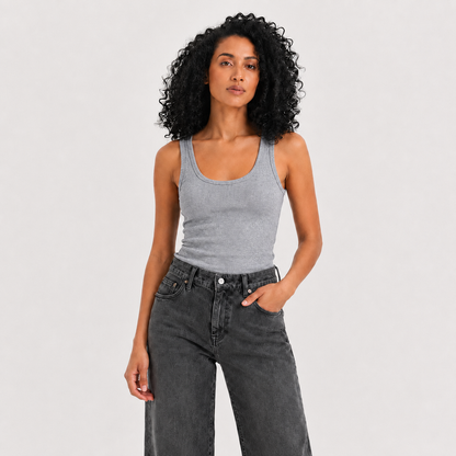 A woman with curly black hair wearing a light gray ribbed tank top and dark gray high-waisted jeans, standing against a light gray studio background.