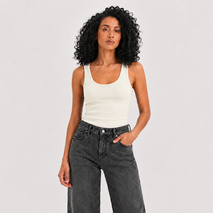 A woman with curly black hair wearing a fitted off-white ribbed tank top and dark gray high-waisted wide-leg jeans, standing against a light gray studio background.