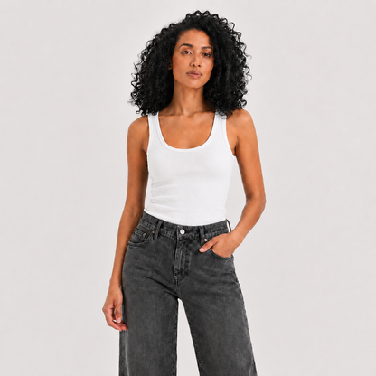 A woman with curly black hair wearing a white ribbed tank top and dark gray jeans, standing against a light gray studio background.
