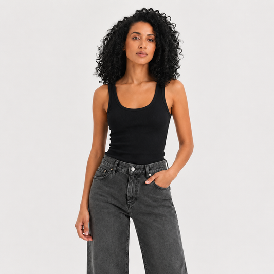 A woman with curly black hair wearing a black scoop-neck tank top and dark gray high-waisted jeans, standing against a light gray studio background.