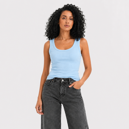 A woman with curly black hair wearing a light blue ribbed tank top and dark gray jeans, standing against a light gray studio background.