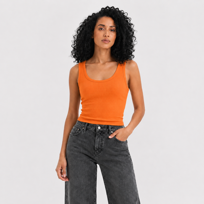 A woman with curly black hair wearing a bright orange ribbed tank top and dark gray jeans, standing against a light gray studio background.