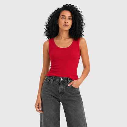 A woman with curly black hair wearing a bright red ribbed tank top and dark gray jeans, standing against a light gray studio background.