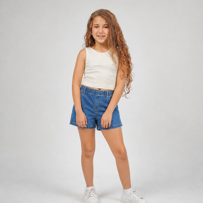A young girl with long, wavy light brown hair stands against a light gray studio background, wearing a sleeveless cream top, blue denim shorts, and white sneakers.