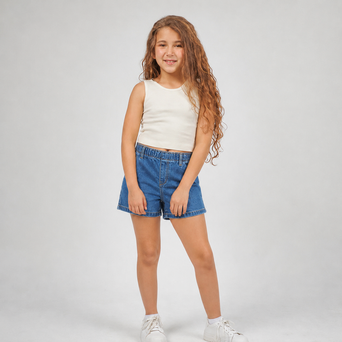 A young girl with long, wavy light brown hair stands against a light gray studio background, wearing a sleeveless cream top, blue denim shorts, and white sneakers.