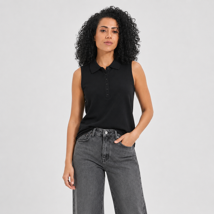 A woman with curly black hair wearing a black sleeveless polo shirt and gray jeans, standing against a light gray studio background.