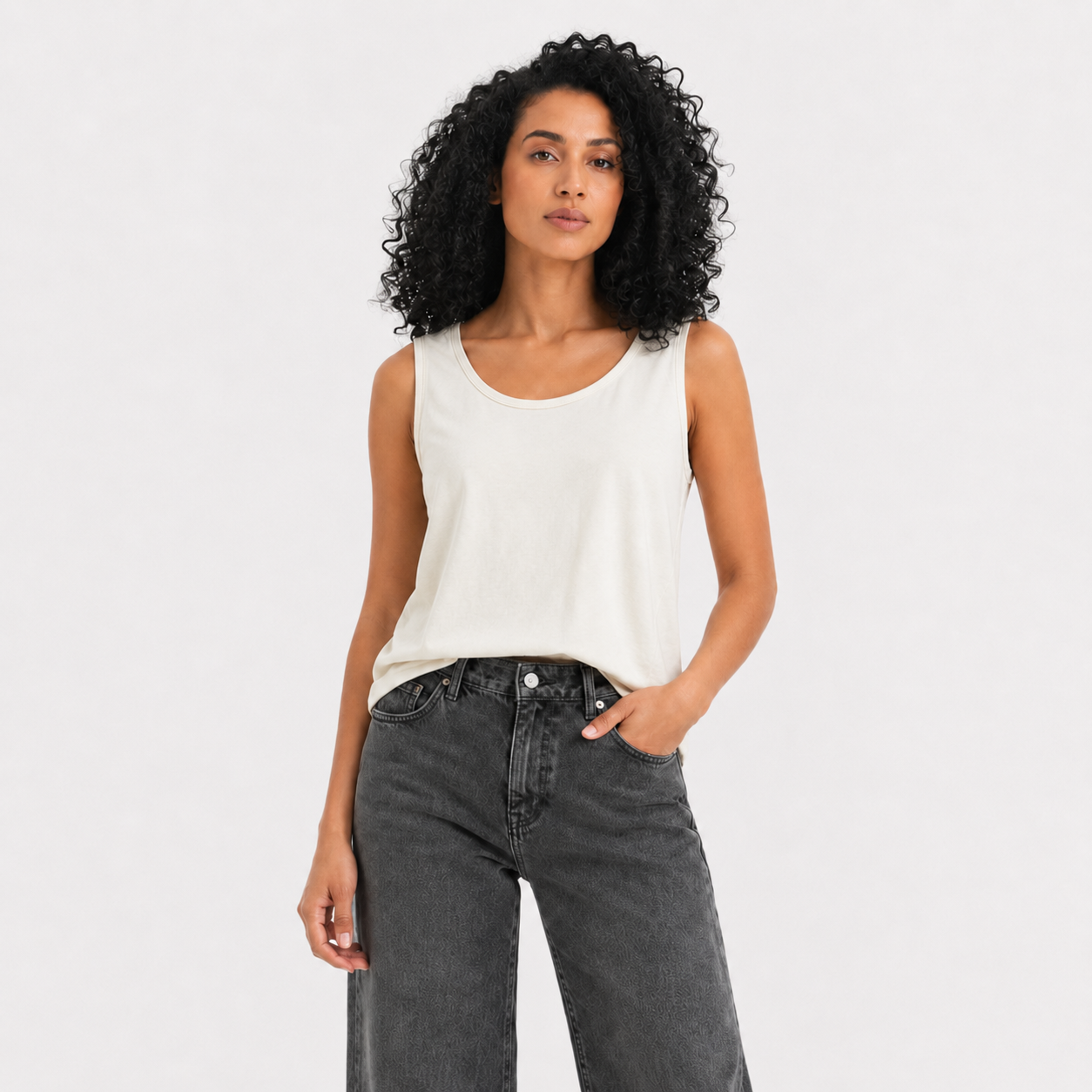 A woman with curly black hair wearing a white sleeveless tank top and dark gray jeans, standing against a light gray studio background.