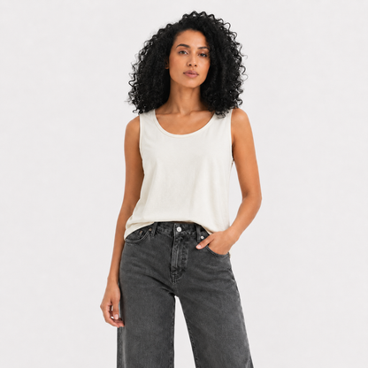 A woman with curly black hair wearing a white sleeveless tank top and dark gray jeans, standing against a light gray studio background.
