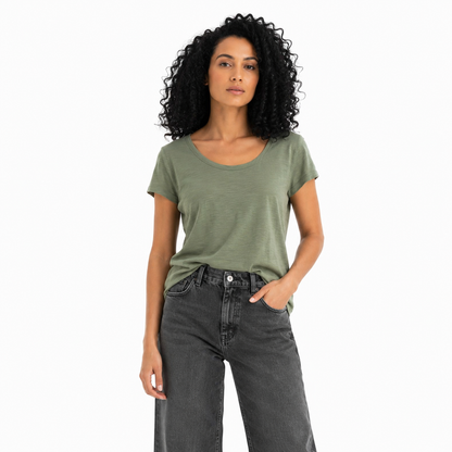 A woman with curly black hair wearing a muted sage green short-sleeve scoop-neck t-shirt and dark gray wide-leg jeans, standing against a plain white background.