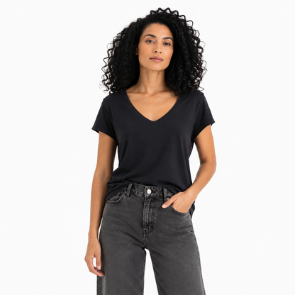 A woman with curly black hair wearing a black short-sleeve V-neck t-shirt and dark gray jeans, standing against a plain white studio background.