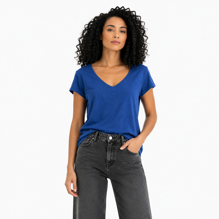 A woman with curly black hair wearing a vibrant royal blue v-neck t-shirt and dark gray jeans, standing against a plain white studio background.