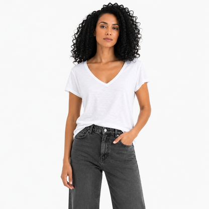 A woman with curly black hair wearing a white short-sleeve V-neck t-shirt and dark gray jeans, standing against a plain white background.