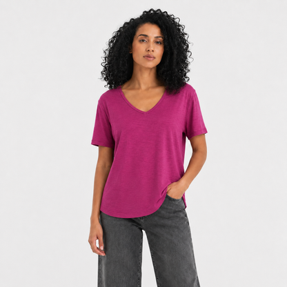 A woman with curly black hair wearing a bright magenta V-neck short-sleeve t-shirt and dark gray jeans, standing against a light gray studio background.