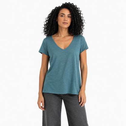 A woman with curly black hair wearing a slate blue v-neck t-shirt and dark gray jeans, standing against a plain white studio background.