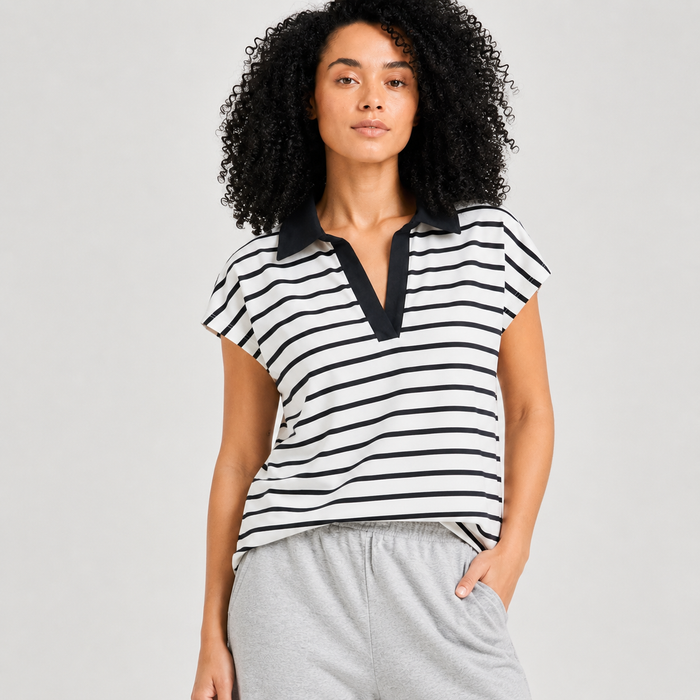 A woman with curly black hair wearing a black and white horizontally striped short-sleeve polo shirt with a black collar, paired with light gray sweatpants, against a light gray studio background.