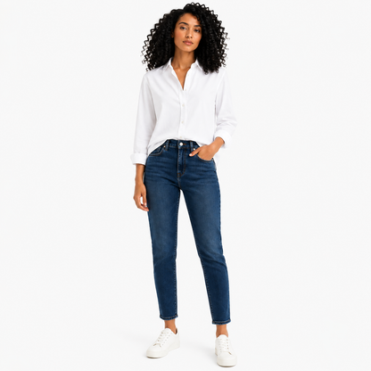 A woman modeling a white long-sleeve button-up shirt and dark blue straight-leg jeans with white sneakers, standing against a plain white studio background.