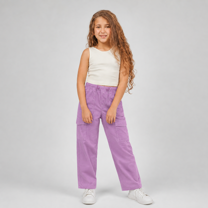 A young girl with long, wavy light brown hair stands against a light gray studio background, wearing lavender purple wide-leg cargo pants, a white sleeveless top, and white sneakers.