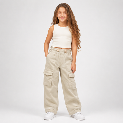A young girl with long wavy brown hair stands against a light gray studio background wearing beige wide-leg cargo pants, a white sleeveless crop top, and white sneakers.