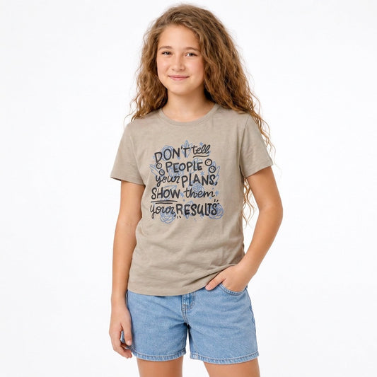 A young girl with long wavy brown hair stands against a white background wearing a dark beige graphic t-shirt and denim shorts. The t-shirt features a dark blue and black typographic design that reads, 'DON'T tell people your plans, show them your results.