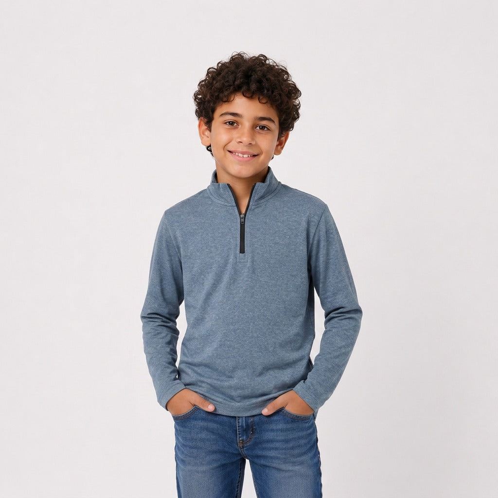 A young boy with curly dark hair wearing a heathered slate blue quarter-zip long-sleeve shirt and blue jeans, standing against a light gray studio background.