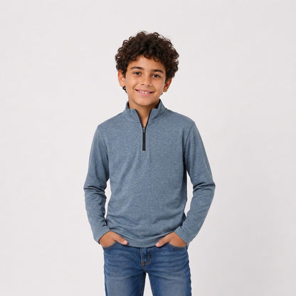 A young boy with curly dark hair wearing a heathered slate blue quarter-zip long-sleeve shirt and blue jeans, standing against a light gray studio background.