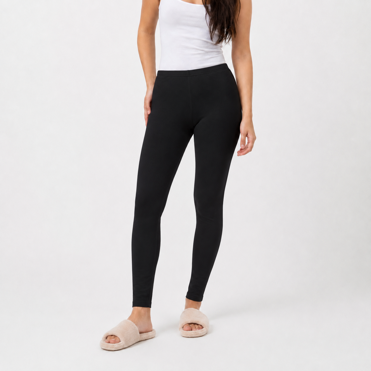 A model wearing black high-waisted leggings, a white tank top, and beige slide sandals, shown from the waist down against a light gray studio background.