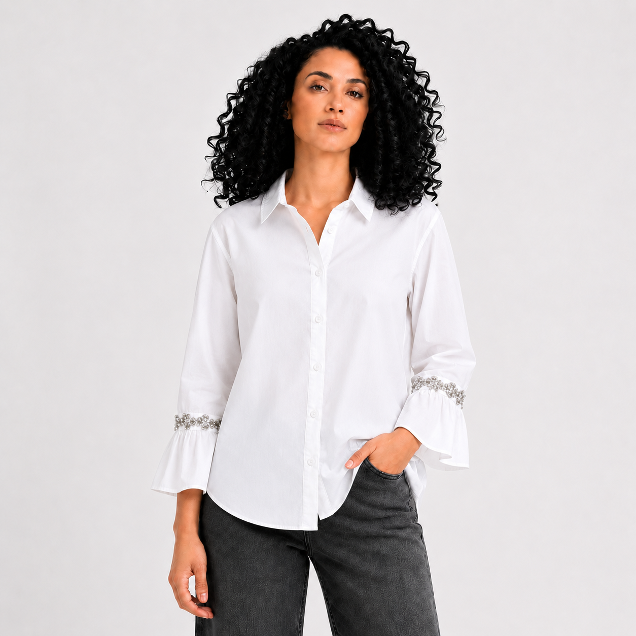 A woman with curly black hair wearing a white long-sleeve button-up blouse with silver beaded cuff details and dark gray jeans, standing against a light gray studio background.