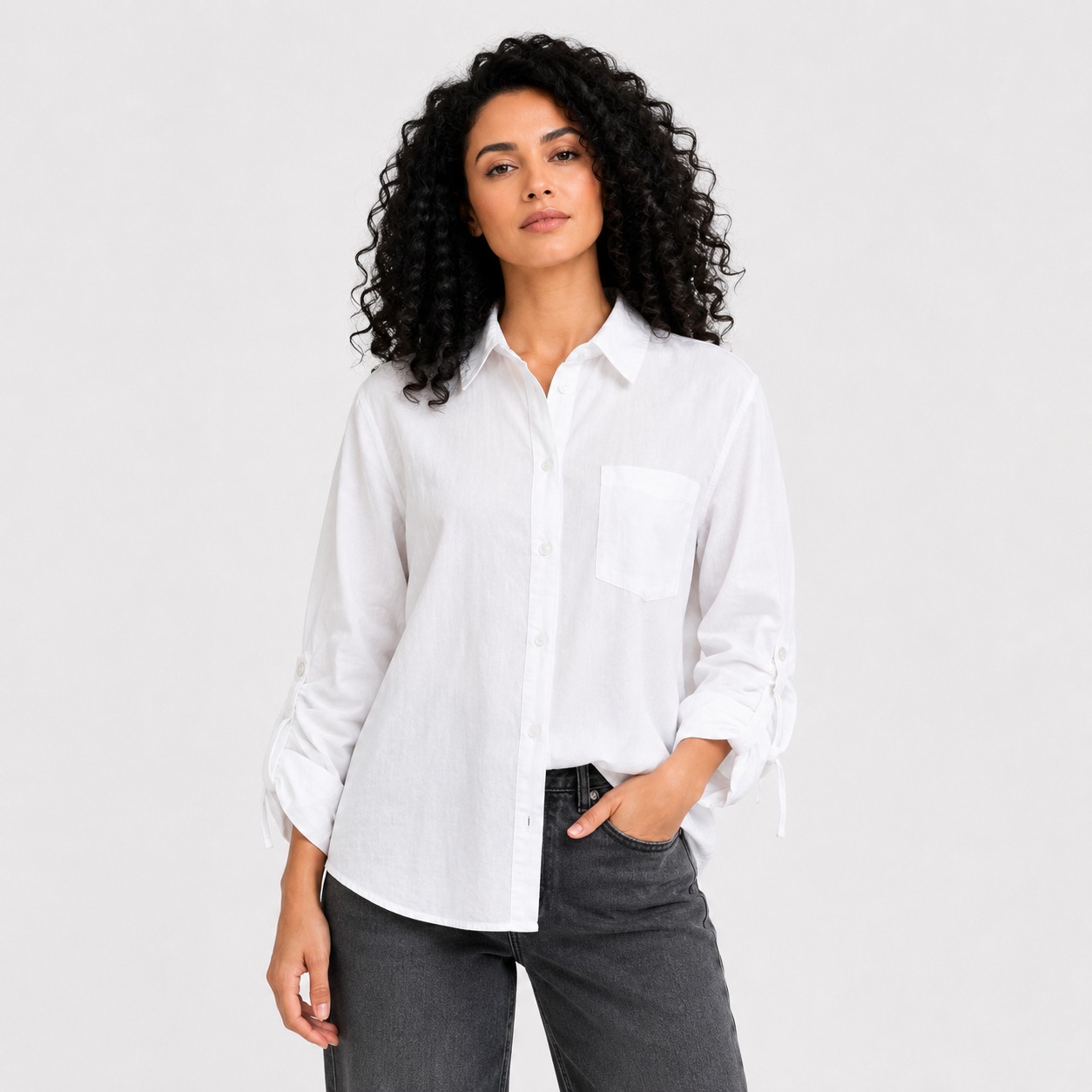 A woman with curly black hair wearing a white linen long-sleeve button-up shirt with rolled cuffs and dark gray jeans, standing against a light gray studio background.