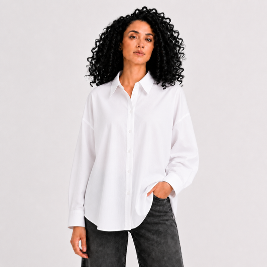A woman with curly black hair wearing a white button-down shirt and dark gray jeans, standing against a light gray studio background.