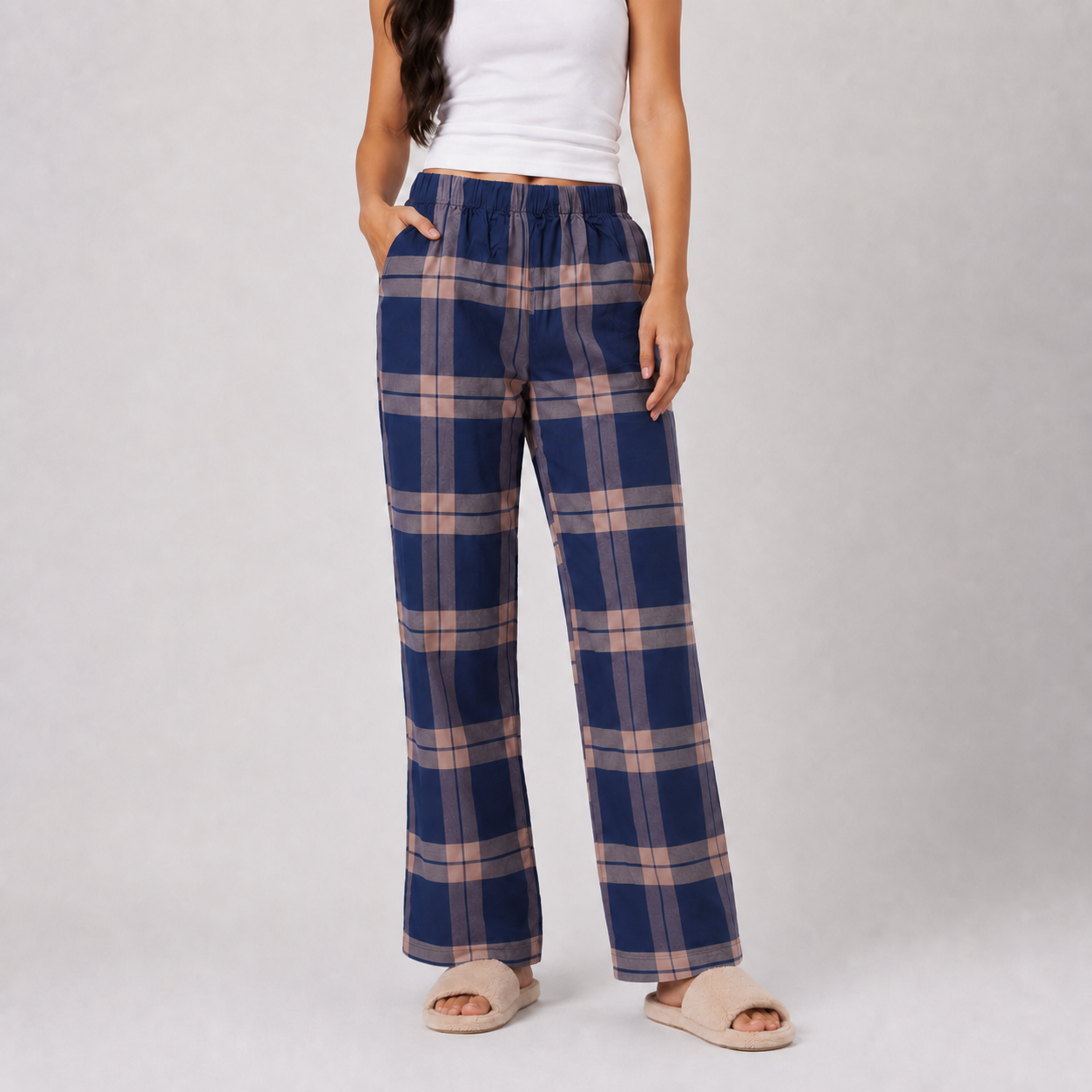 A model wearing blue and beige plaid wide-leg lounge pants, paired with a white tank top and beige slide sandals, standing against a light gray studio background.