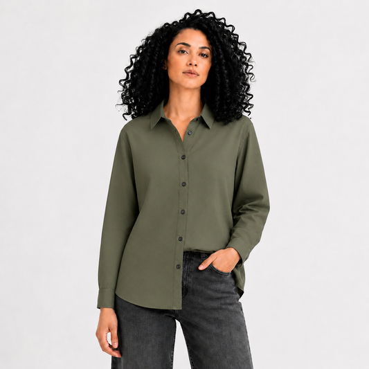 A woman with curly black hair wearing an olive green long-sleeve button-up shirt and dark gray jeans, standing against a light gray studio background.