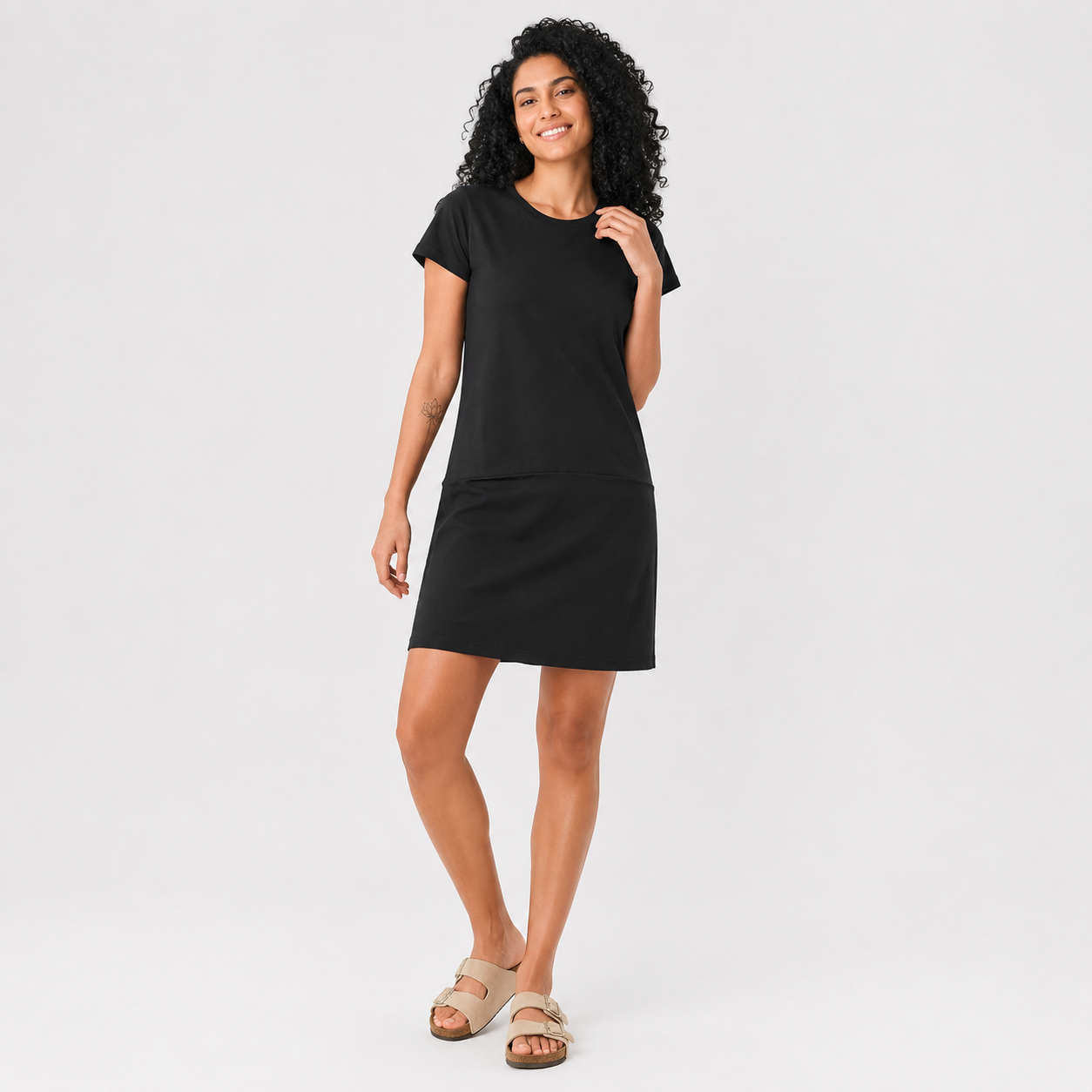 A woman modeling a black short-sleeve sleeping dress with side pockets, paired with beige slide sandals, against a light gray studio background.