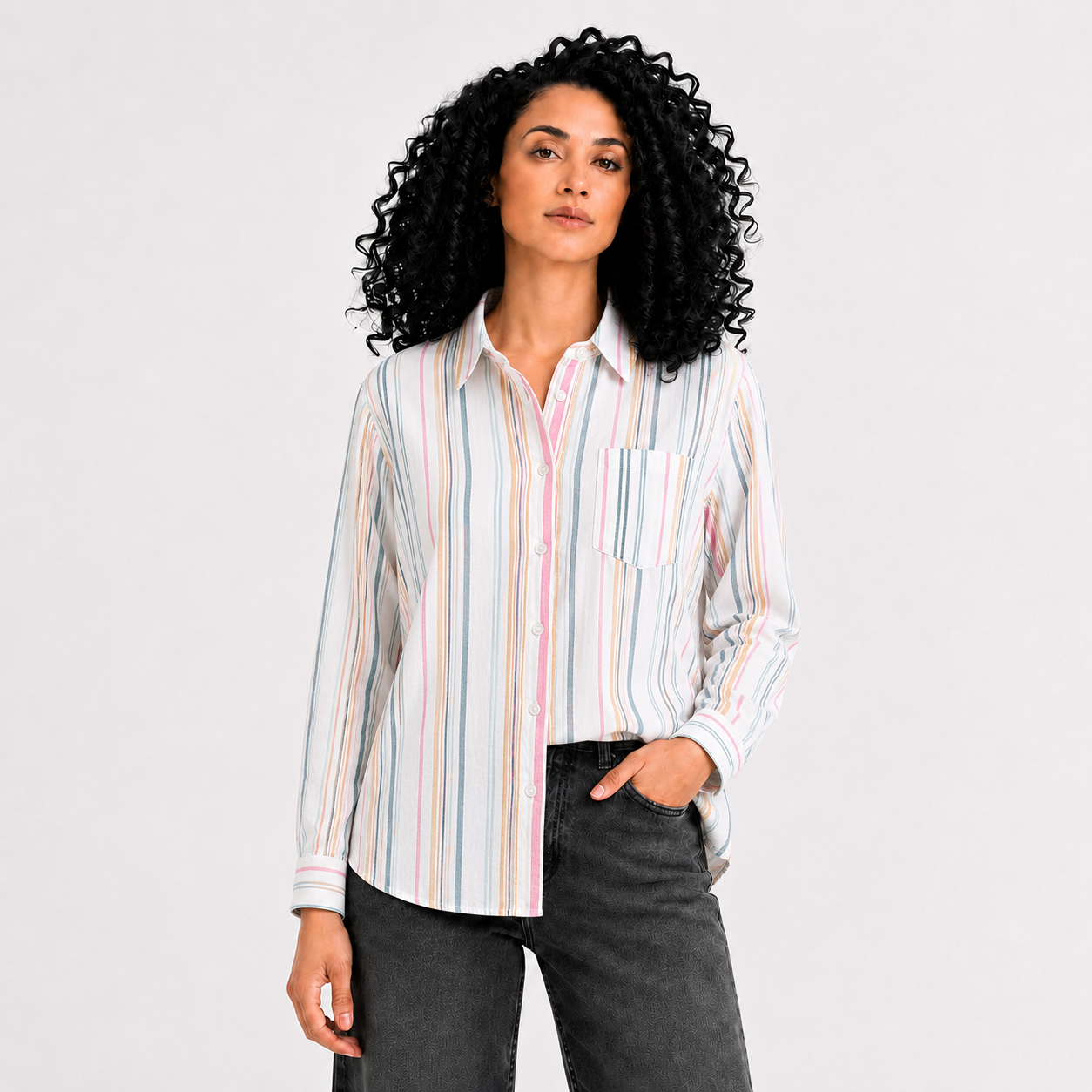 A woman with curly black hair wearing a long-sleeve button-up shirt with vertical pastel stripes in pink, blue, yellow, and white, paired with dark gray jeans, against a light gray studio background.