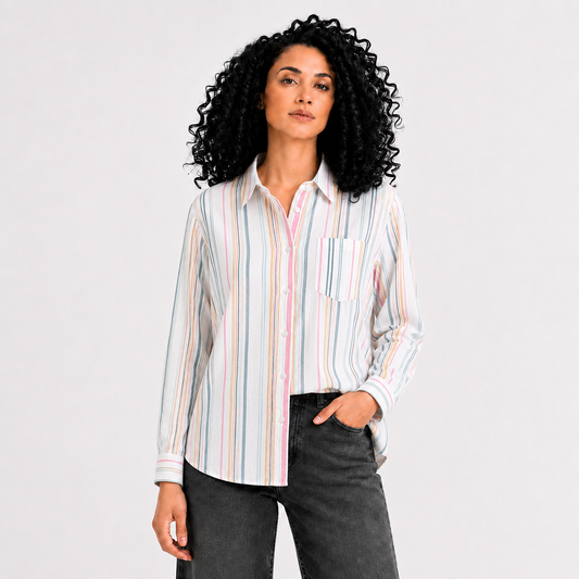 A woman with curly black hair wearing a long-sleeve button-up shirt with vertical pastel stripes in pink, blue, yellow, and white, paired with dark gray jeans, against a light gray studio background.