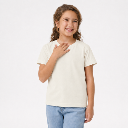 A young girl with curly brown hair smiling and posing in a plain cream-colored short-sleeve t-shirt and light blue jeans against a solid white studio background.
