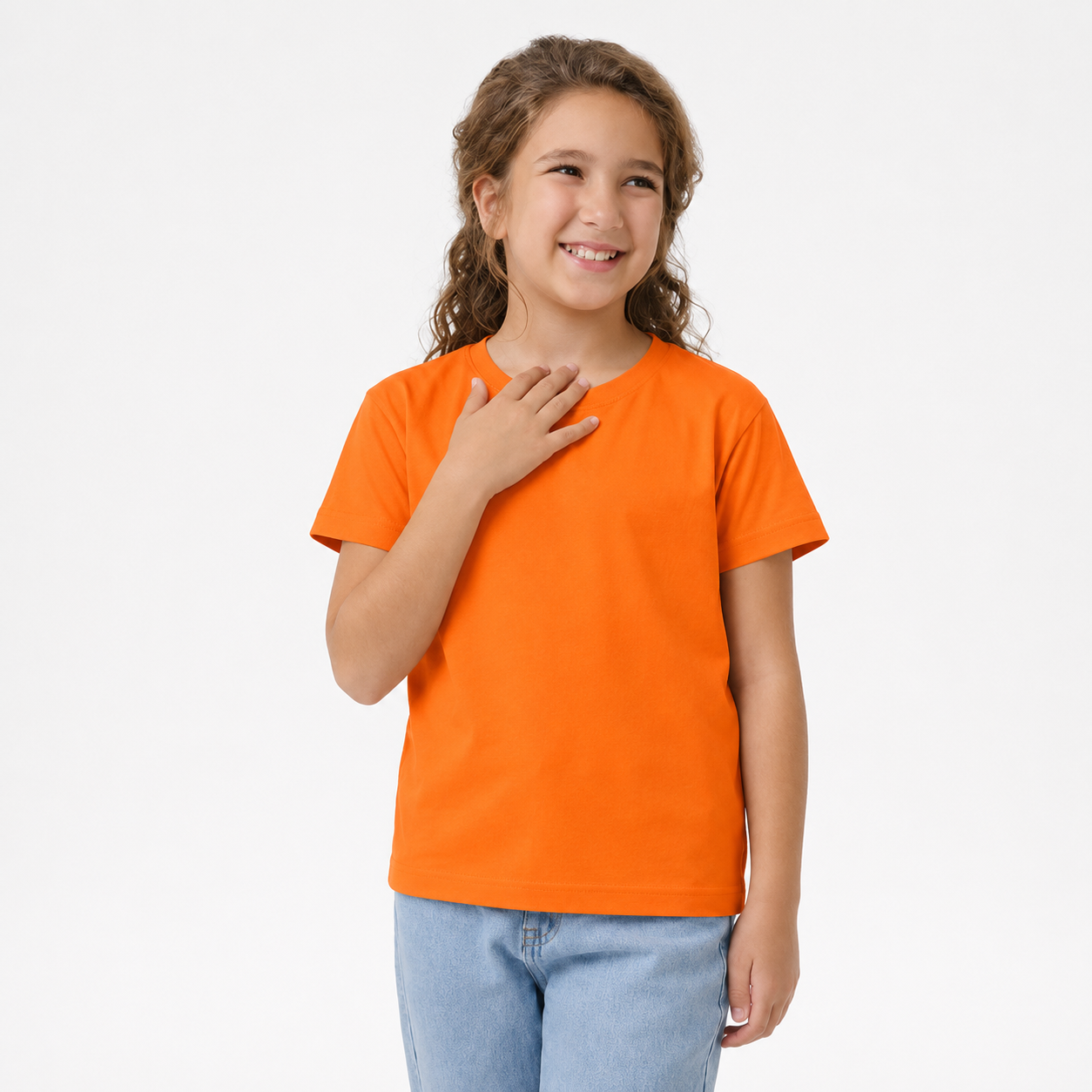 A young girl with curly brown hair smiling and posing in a bright orange short-sleeve t-shirt and light blue jeans against a solid white studio background.